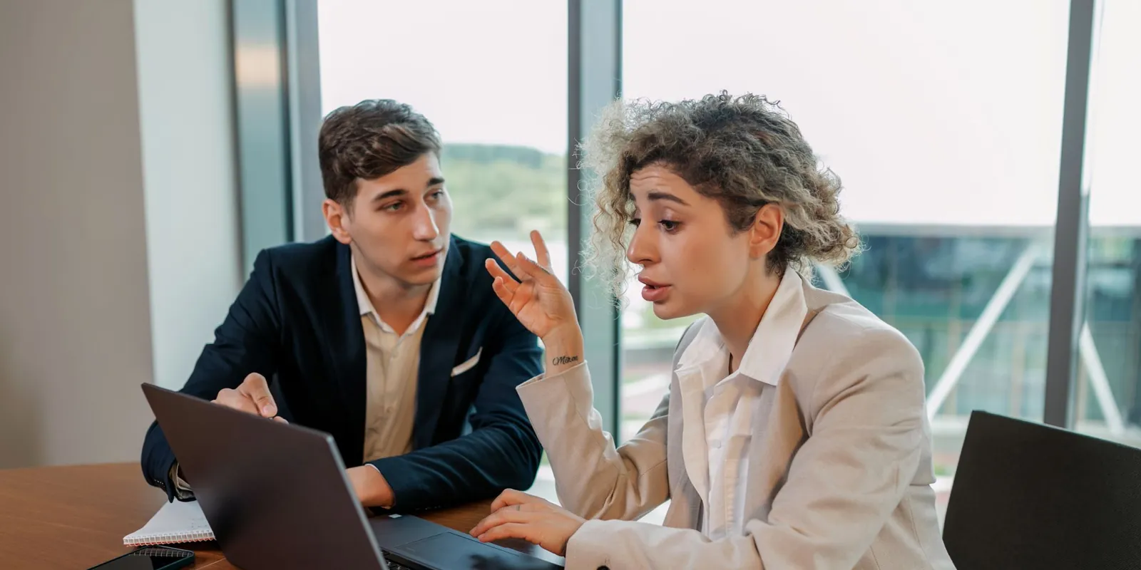 Two colleagues discussing a project at a table with a laptop in a bright office