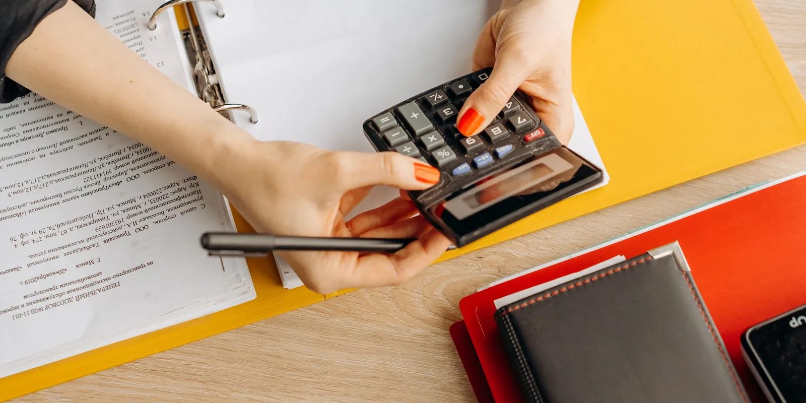 Hands using a calculator over financial paperwork and a binder on a desk