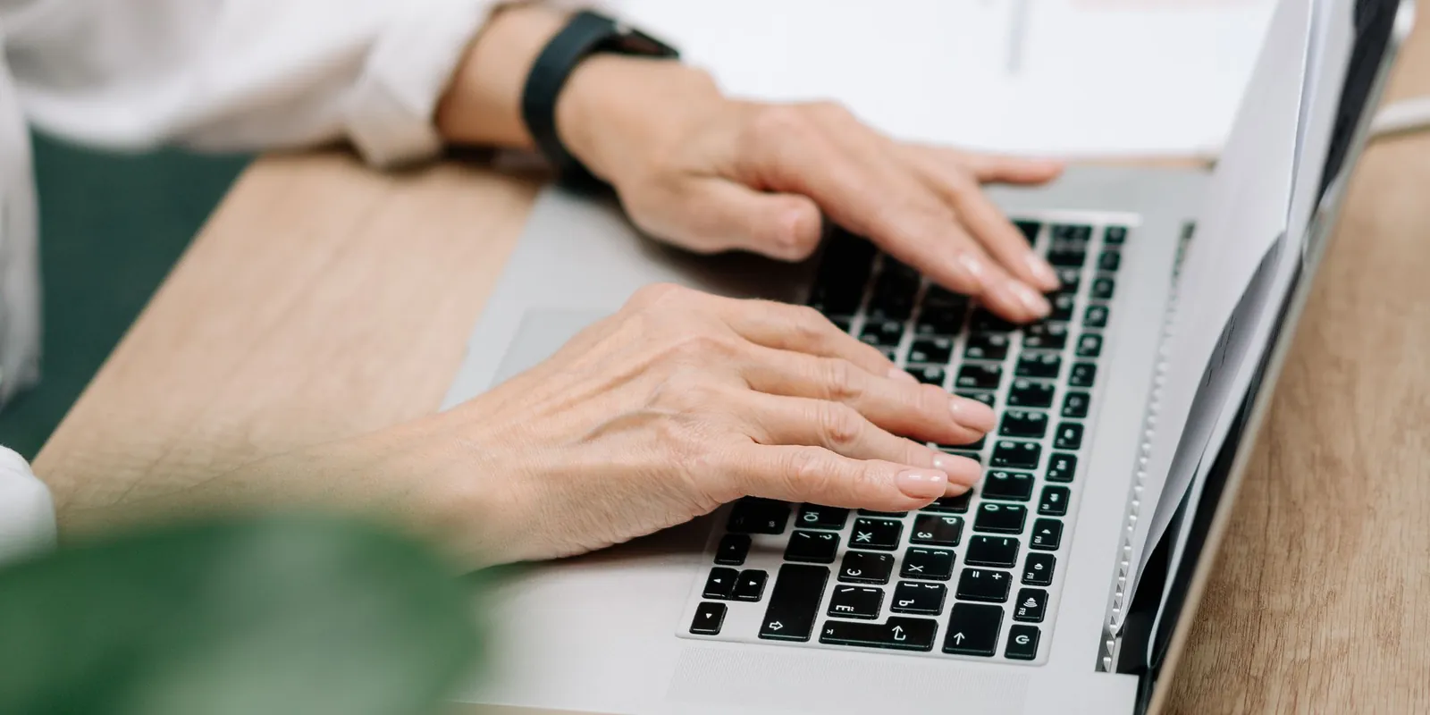 Hands typing an email on a laptop at a wooden desk beside papers and a plant